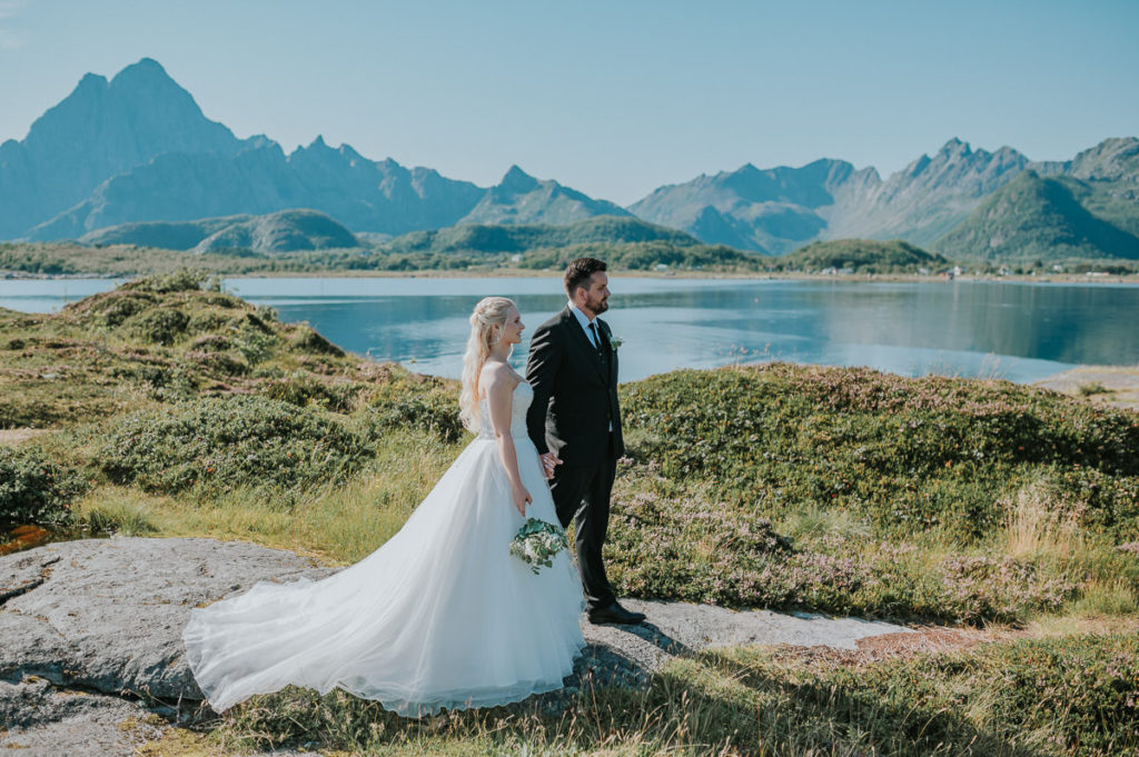 Bridal couple enjoying a sunny and warm summer day while watching the sea and mountains in Lofoten on the day of their adventure wedding in Nyvågar Kabelvåg