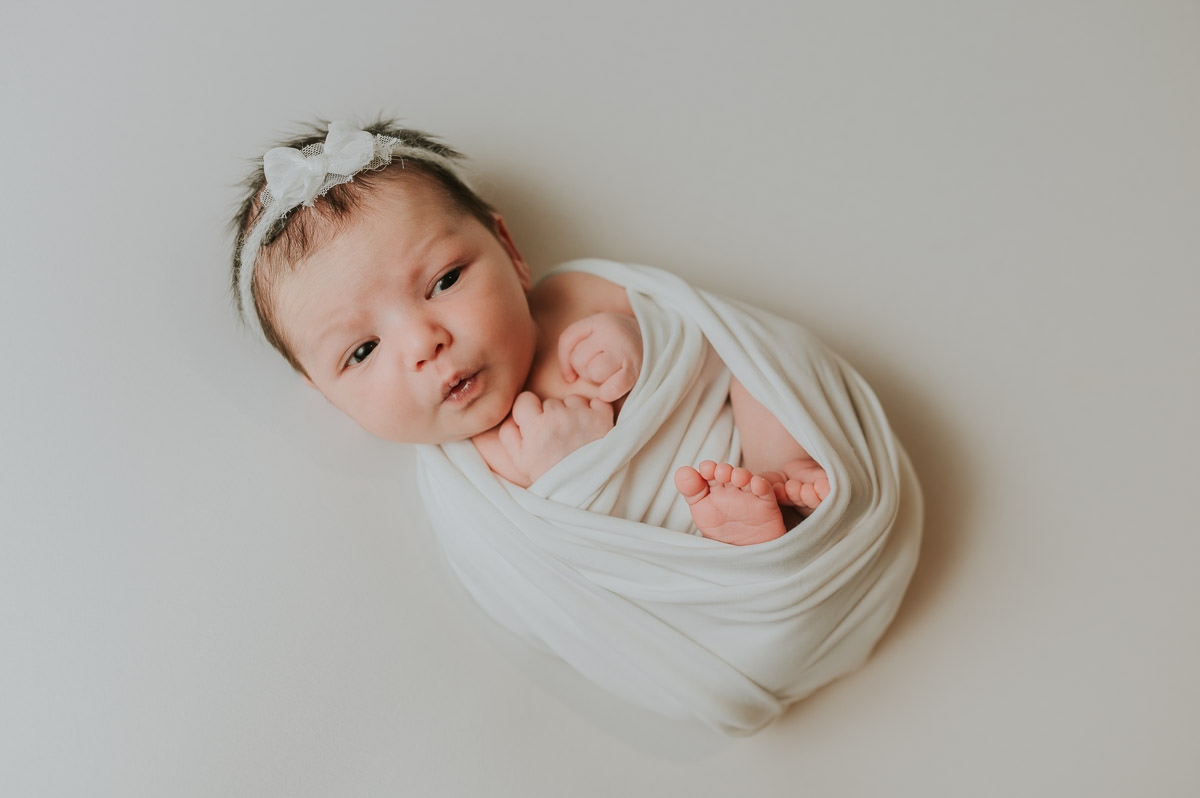 Nyfødt jente med masse hår og hvit hårbånd ligger på hvit fotobakgrunn på nyfødtfotografering i Alta hos TS Foto Design