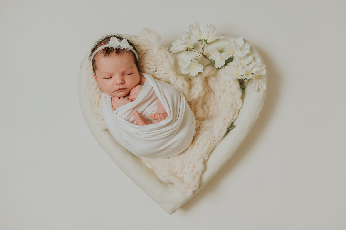 Nyfødt jente med masse hår og hvit hårbånd ligger i et hvit hjerte fotoprop med hvit fotobakgrunn på nyfødtfotografering i Alta hos TS Foto Design