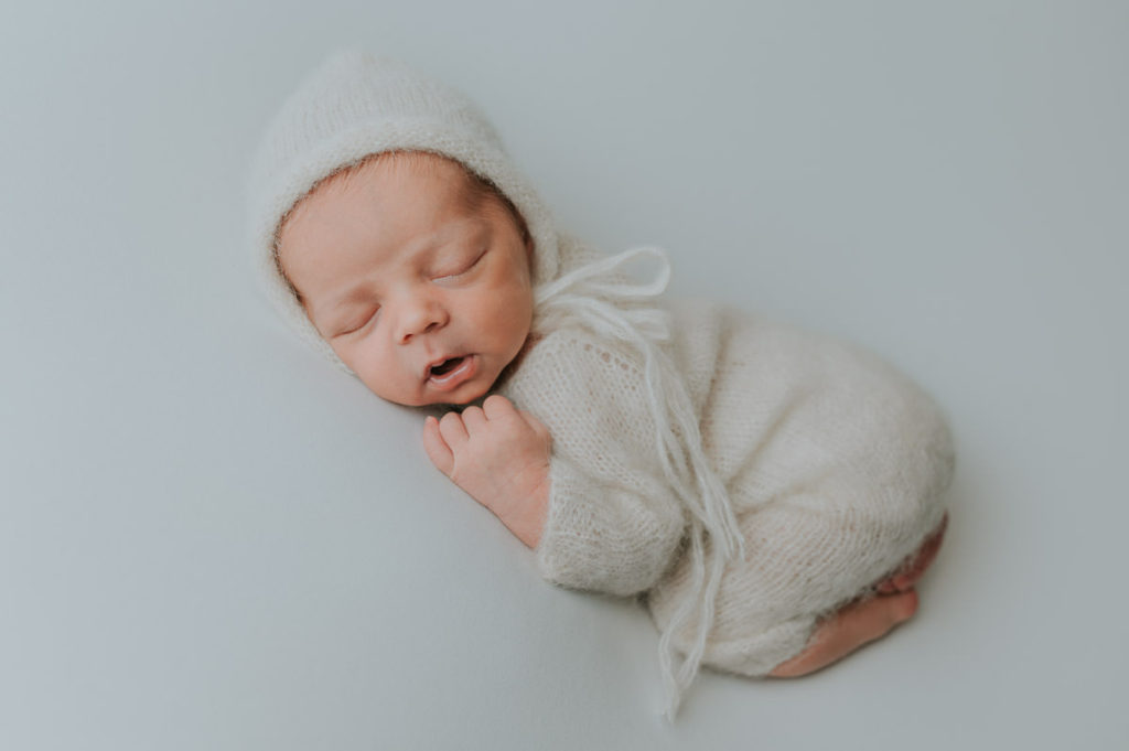 Nyfødt gutt i hvit strikket drakt og lue sover på hvit fotobakgrunn på nyfødtfotografering i Alta hos TS Foto Design