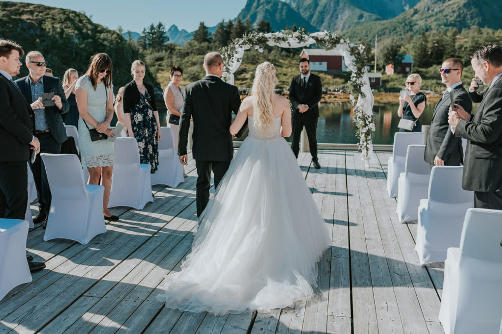 Outdoor summer wedding ceremony at Nyvågar rorbu hotel in Kabelvåg Lofoten. The groom is waiting for his bride while the father walks the bride down the aisle