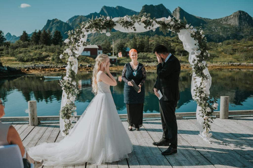 Bride and groom reading their vows to each other on the day of their adventure wedding in Lofoten. The ceremony is held on a pier in Lofoten oevrlooking stunning mountains and the sea. Both bride and groom have tears in their eyes and they are wiping them with the hands