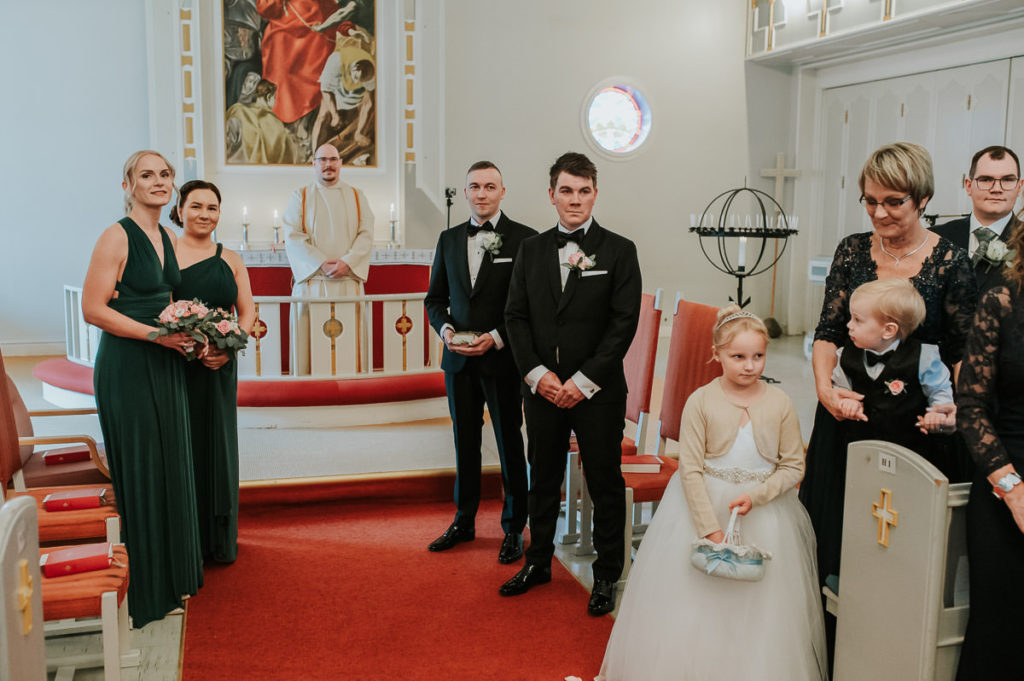 Brudgommen, forlovere og foreldre venter på bruden som spaserer til alteret sammen med sin far