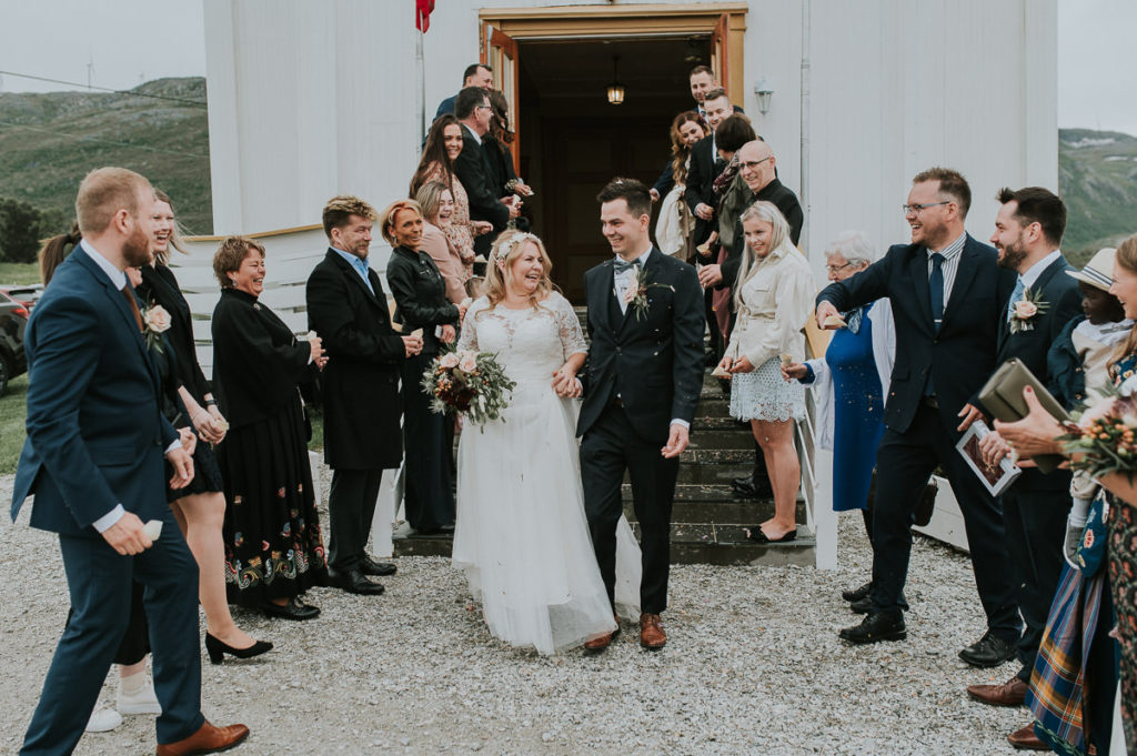 Bride and groom walking out of Hillesøy church after their wedding ceremony. The guests are throwing fresh flower petal confetti at them and the couple is laughing - captured by Tromsø wedding photographer TS Foto Design