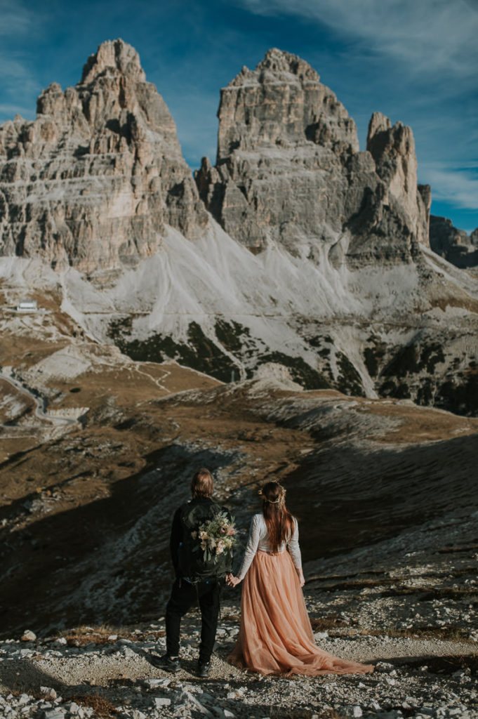 Bride and groom facing spectacular mountains in Italy with a rocksack and bridal bouquet in it - elopement in Dolomites