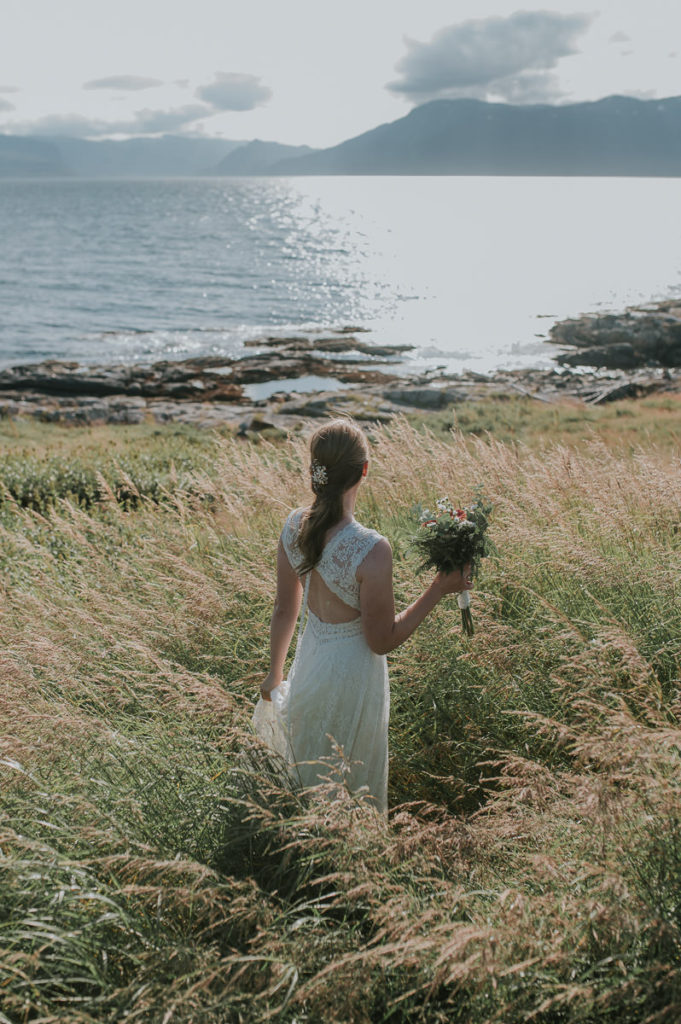 Bohemian bride in a lace dress with open back standing in a golden grass field watching a sea view and mountains on a sunny day in Norway