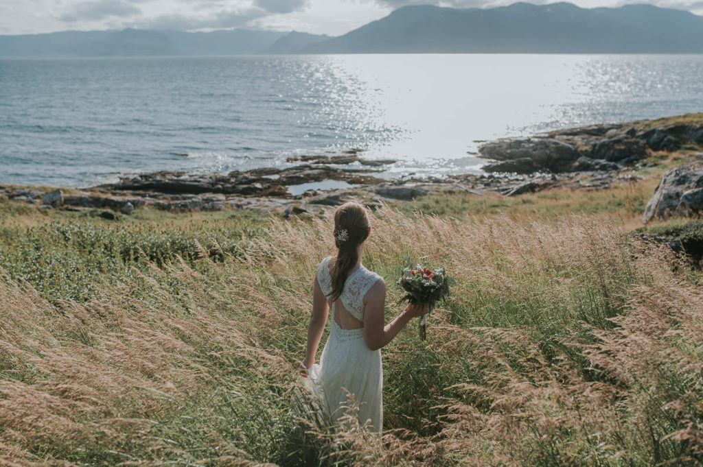 Bohemian bride in a lace dress with open back standing in a golden grass field watching a sea view and mountains on a sunny day in Norway