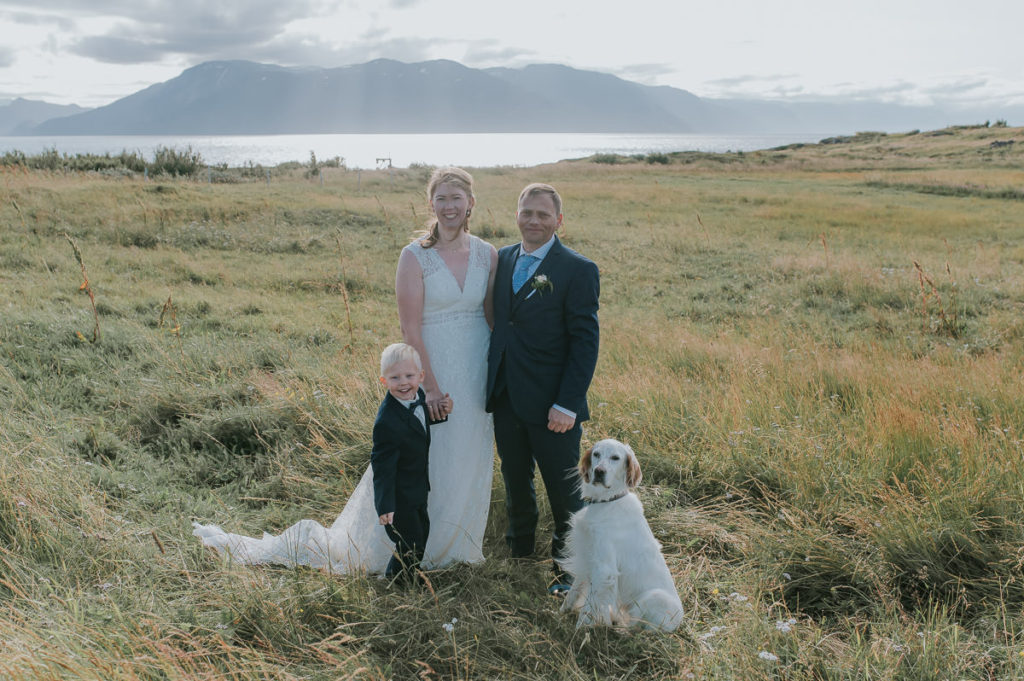 Bride and groom with their son and dog in a grass field with a sea and mountains behind them