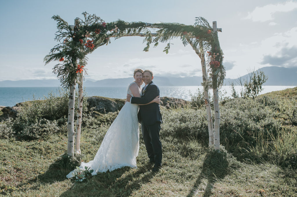Bride and groom under a floral arch with a sea and mountain view on a day of their outdoor intimate wedding in Alta Norway