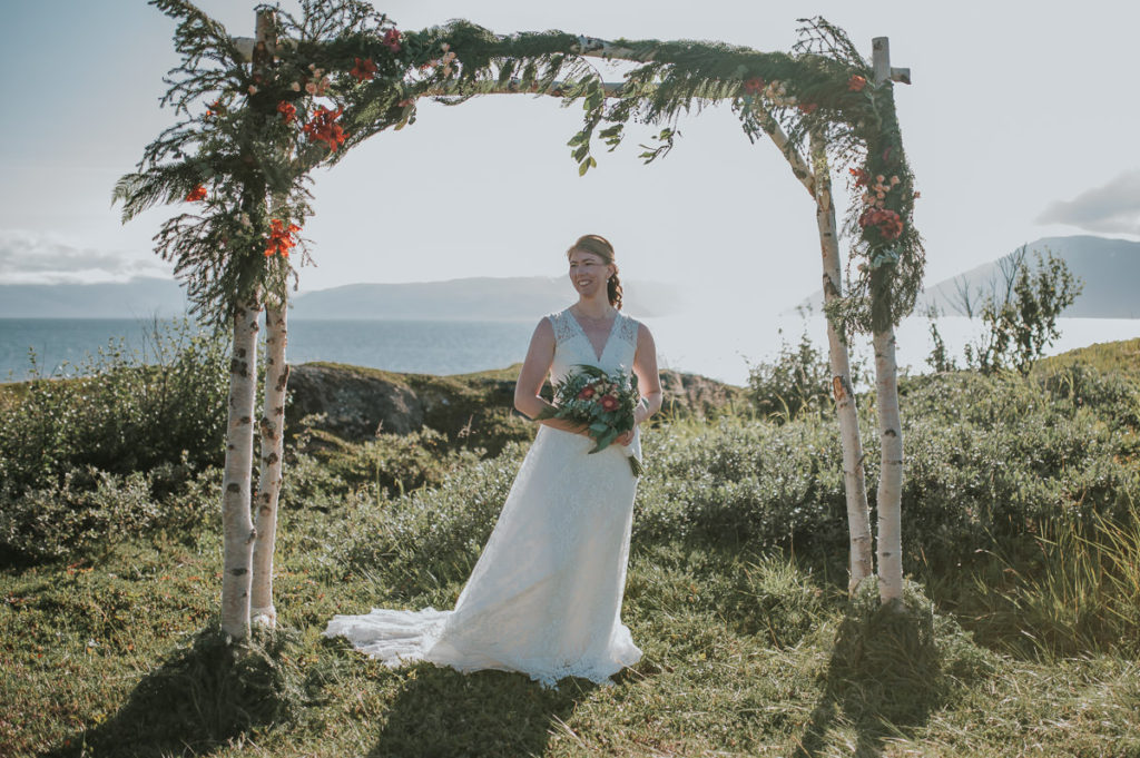 Bride under a floral arch with a sea and mountain view on a day of their outdoor intimate wedding in Alta Norway