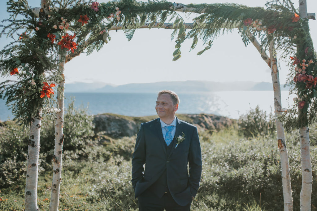 Groom under a floral arch with a sea and mountain view on a day of their outdoor intimate wedding in Alta Norway