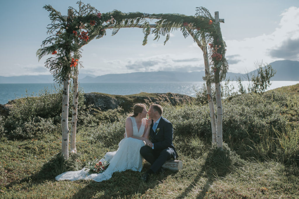 Bride and groom under a floral arch with a sea and mountain view on a day of their outdoor intimate wedding in Alta Norway
