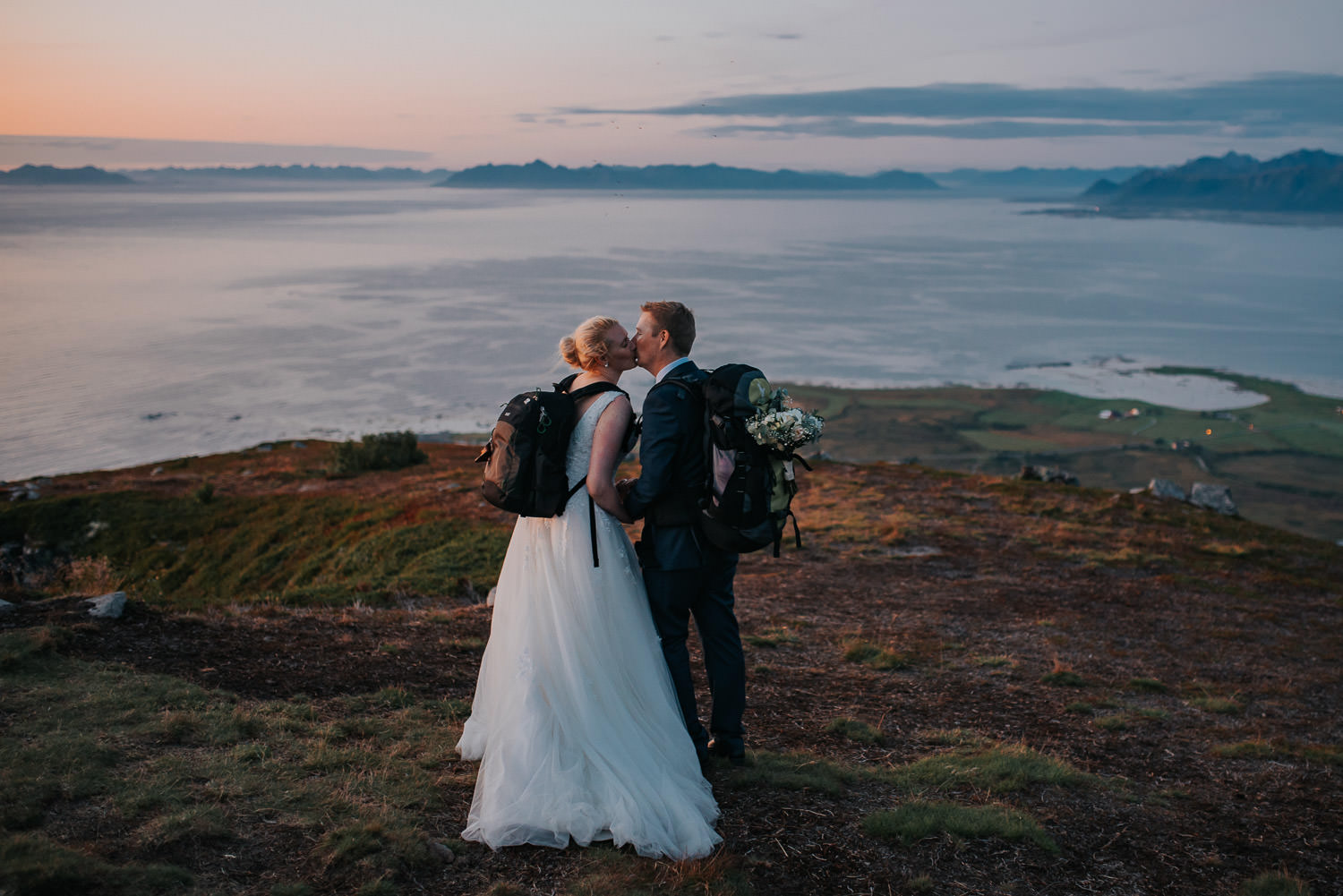 Bride and groom in the mountains of Lofoten in Norway with their backpacks and flowers tucked in the backpack kissing each other at the end of their elopement day