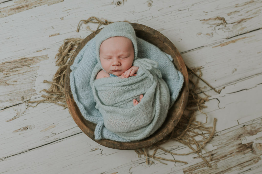 Nyfødtgutt inntullet i lyseblå sjal sover i en rund trebolle på nyfødtfoto i fotostudio i Alta