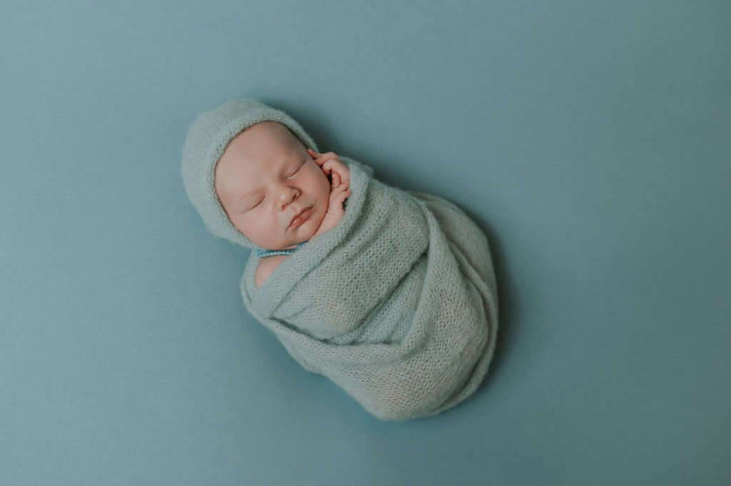 Nyfødtgutt inntullet i lyseblå sjal sover på blå fotobakgrunn på nyfødtfoto i fotostudio i Alta