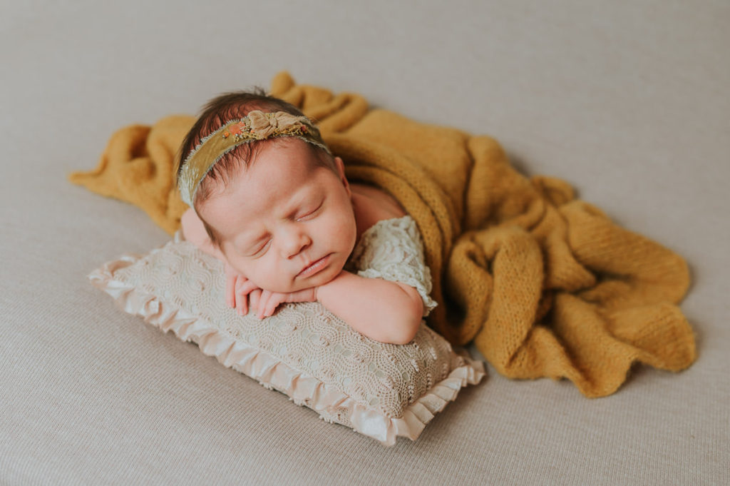 Nyfødt jente sover søtt på en beige fotobakgrunn med okergule toner på hårbånd og teppe
