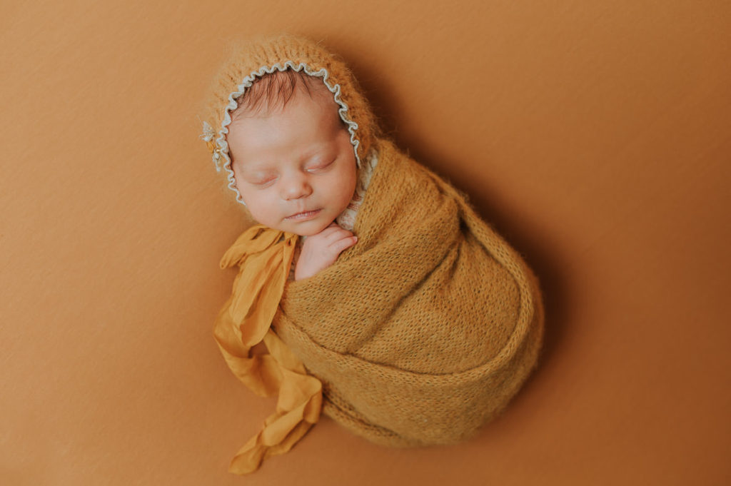 Nyfødtjente med en gul strikket lua inntullet i en sjal på okergul fotobakgrunn 