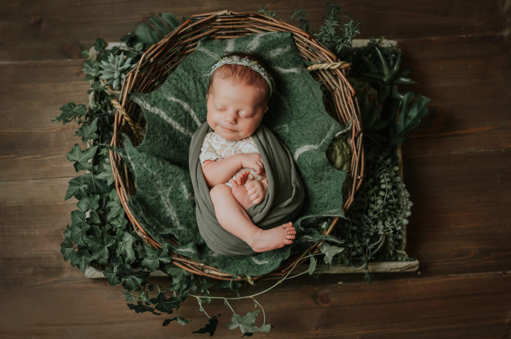 Nydelig baby jente med grønn hårbånd ligger i en kurv blant grønne planter på nyfødtfotografering i Alta hos TS Foto Design