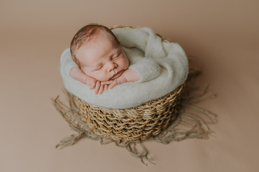 Nyfødt gutt i en hvit ulldrakt soveri en kurv på beige fotobakgrunn i fotostudio i Alta på nyfødtfoto