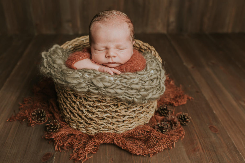 Søt gutt med rustfarget ulldrakt på sover i en kurv på brun fotobakgrunn på nyfødtfoto hos TS Foto Design i Alta