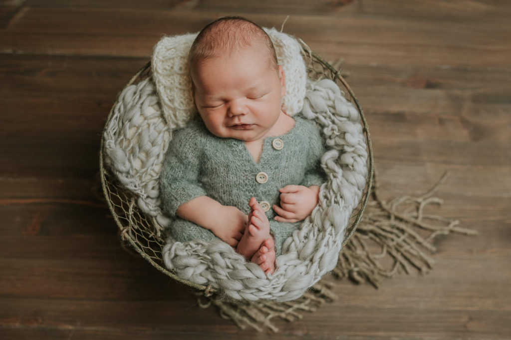 Nyfødt gutt i grønn strikket drakt  sover 
 og smiler i søvne i en kurv på brun bakgrunn