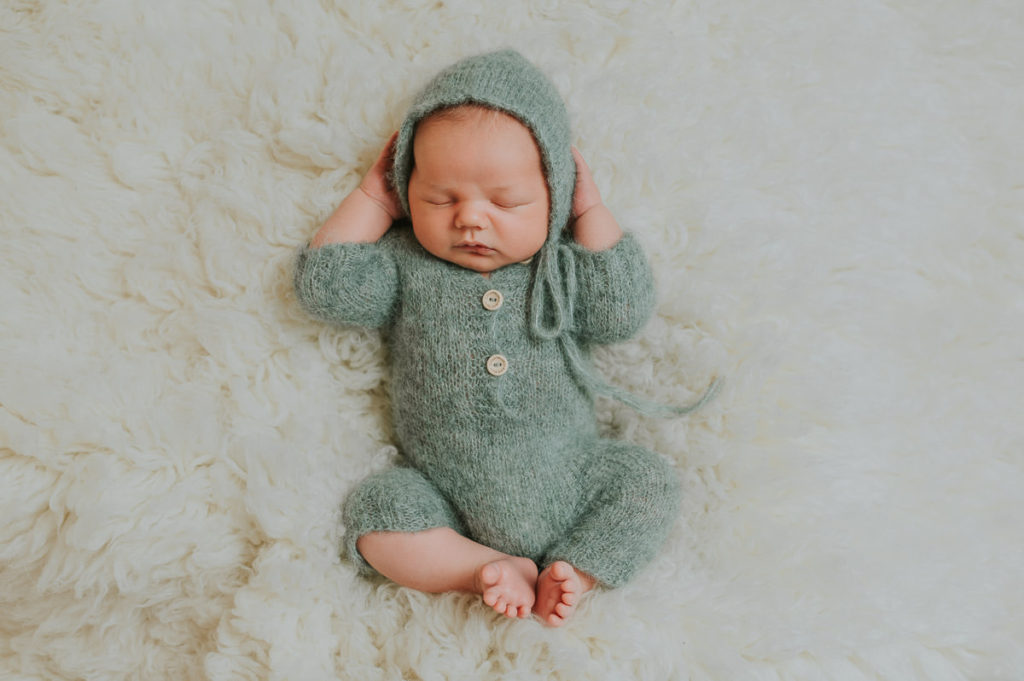 Nyfødt gutt i grønn strikket drakt og lue ligger avslappet på en myk flokati teppe på nyfødtfoto 