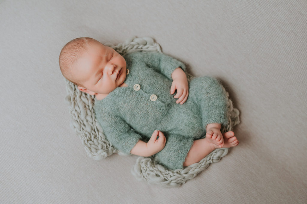 Nyfødt gutt i grønn strikket drakt og lue ligger avslappet på en beige bakgrunn og sover