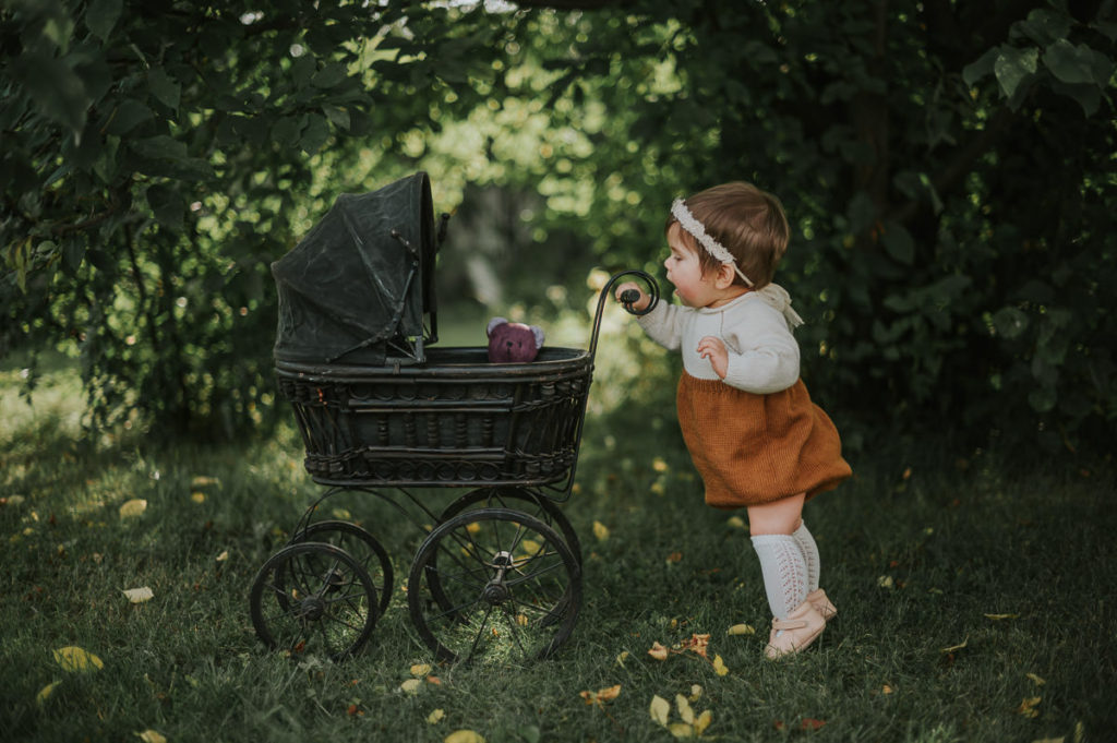 Skjønn ettåring jente triller en vintage dukkevogn blant fine sommer omgivelser i en hage i Alta