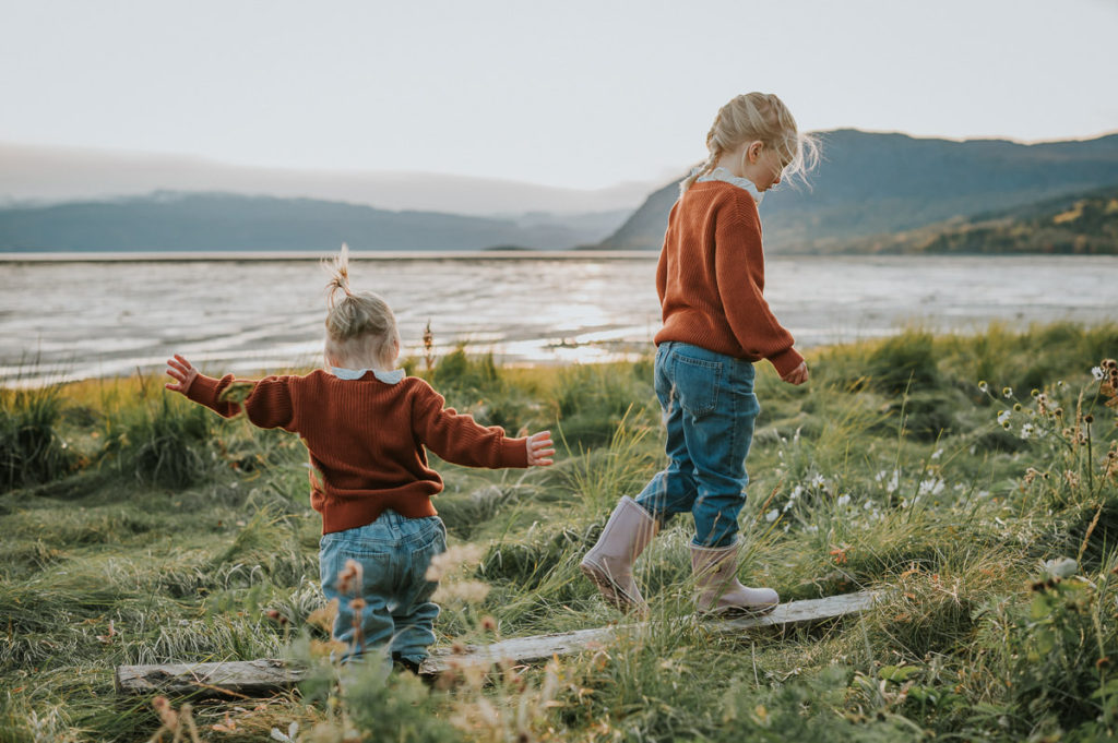 To søstre leker i fjæra i Alta med sjø og fjell i bakgrunn - barnefotografering i Alta