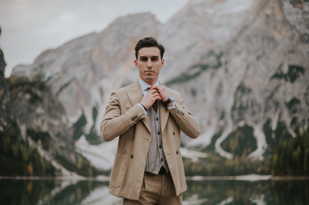 Handsome groom in a beige suite in front of a stunning mountain lake view in Italian Dolomites