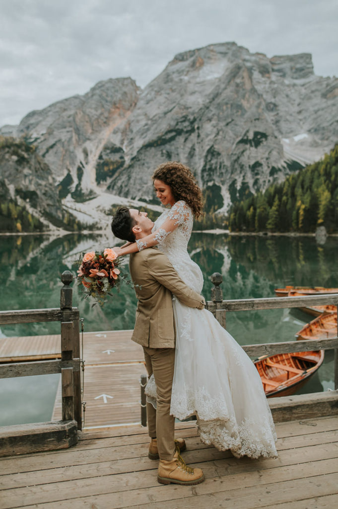 Portrait of a happy couple where the groom lifted his bride and swirls her around while she is laughing, all happens in front of a stunning lake Braies Lago di Braies on the day of their elopement in Italy