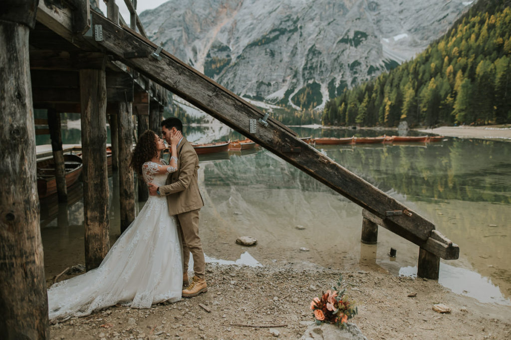 Bride and groom kissing under a boat house next to a mountain lake in Italy