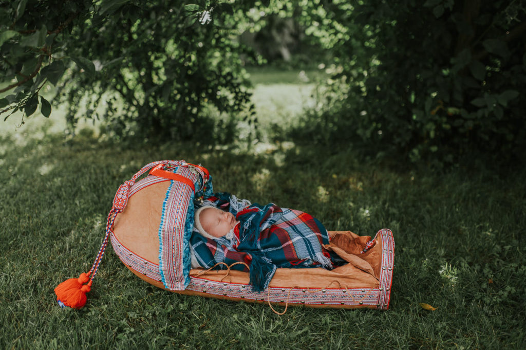 Nyfødtfoto med samisk baby utendørs i Alta i en grønn hage hvor babyen ligger i en håndlaget komsa og sover