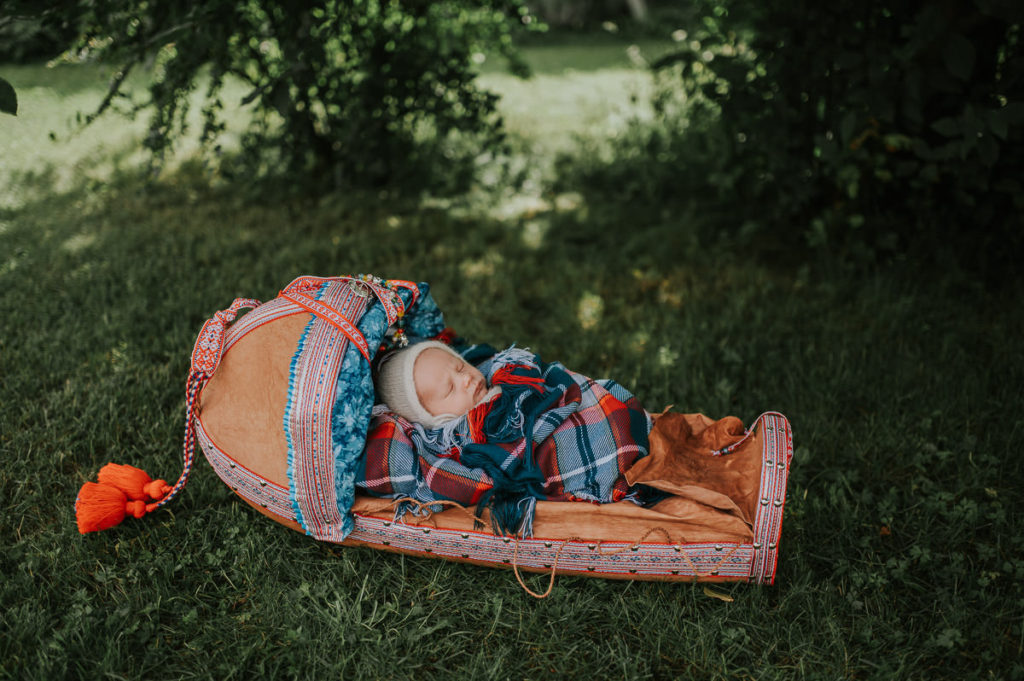 Nyfødtfotografering med samisk baby utendørs i Alta i en grønn hage hvor babyen ligger i en håndlaget komsa og sover