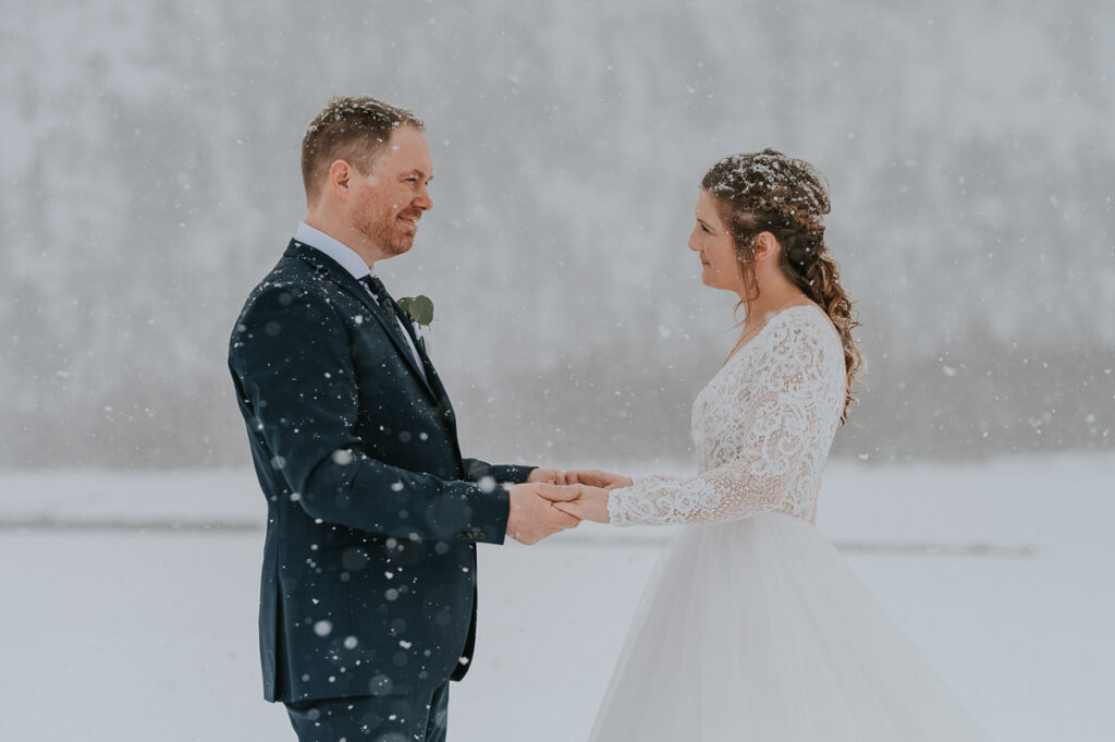 Bride and groom holding hands  in front of a beautiful wonderland  winter landcape in Alta Northern Norway under a beautiful blizzard snowfall right before their dogsledding winter wedding ceremony in Norway