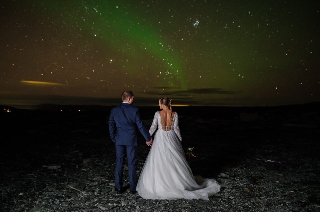 Elopement in Norway under the northern lights. Bride and groom standing in the darkness enjoying beautiful Aurora Borealis display in the sky