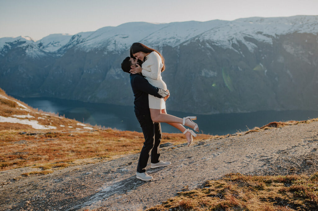 Happy couple on a mountaintop with snowy caped mountains in the background and a fjord view. The guy lifted his girlfriend and both look happy right after the proposal in Aurland Norway