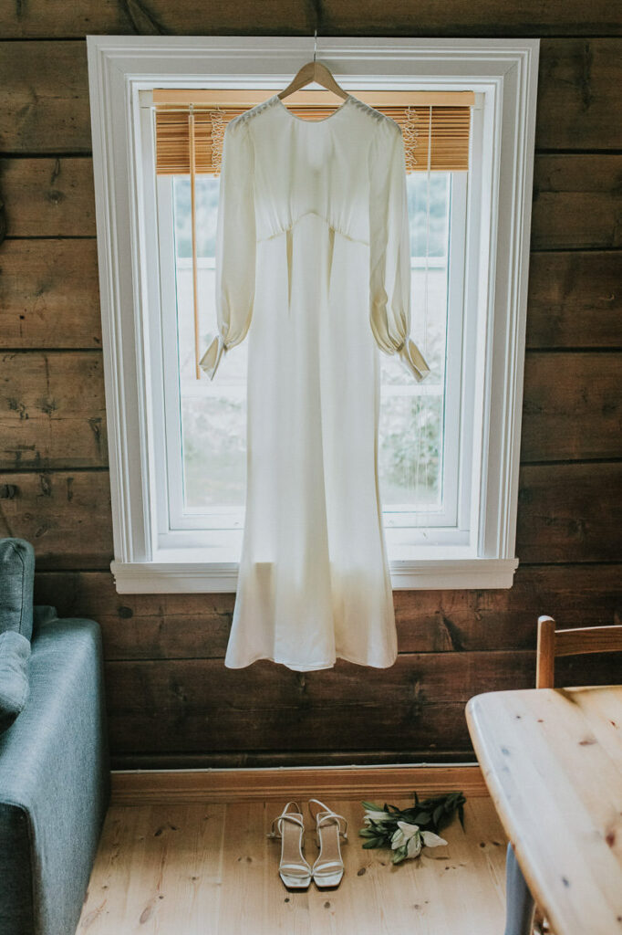 Ivory long sleeve wedding dress hanging in front of window in a cozy cabin with wooden walls