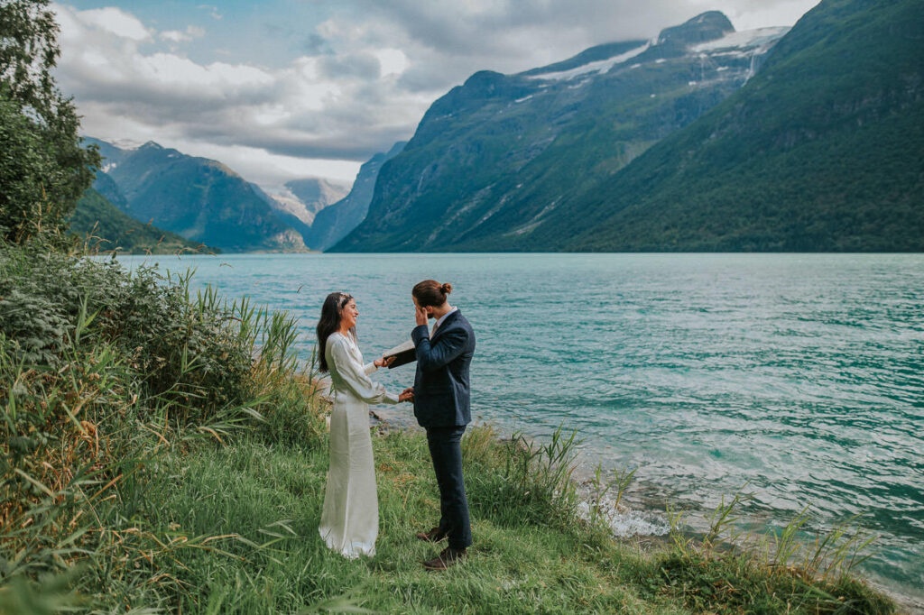 Intimate elopement ceremony by the lake Lovatnet in Loen, Western Norway. The bride is reading her vow from the vow book and groom is drying up some tears