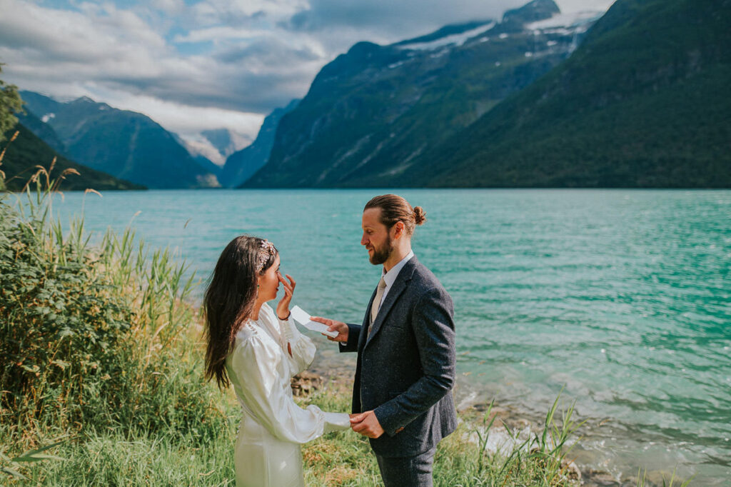 Intimate elopement ceremony by the lake Lovatnet in Loen, Western Norway surrounded by mountains and glaciers. The groom is reading his vow from the vow book and the bride is drying up her tears