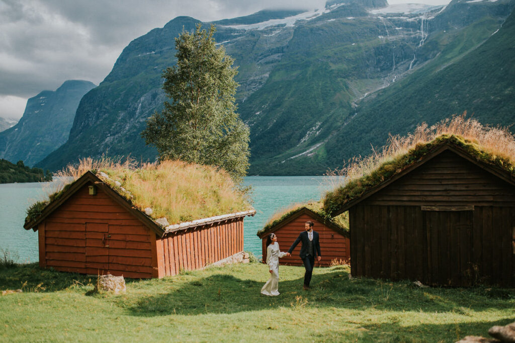 Bride and groom walking among cozy and authentic old cabins with grass growing on the roofs and a gorgeous lake and mountain background - in Loen Western Norway on the day of their elopement