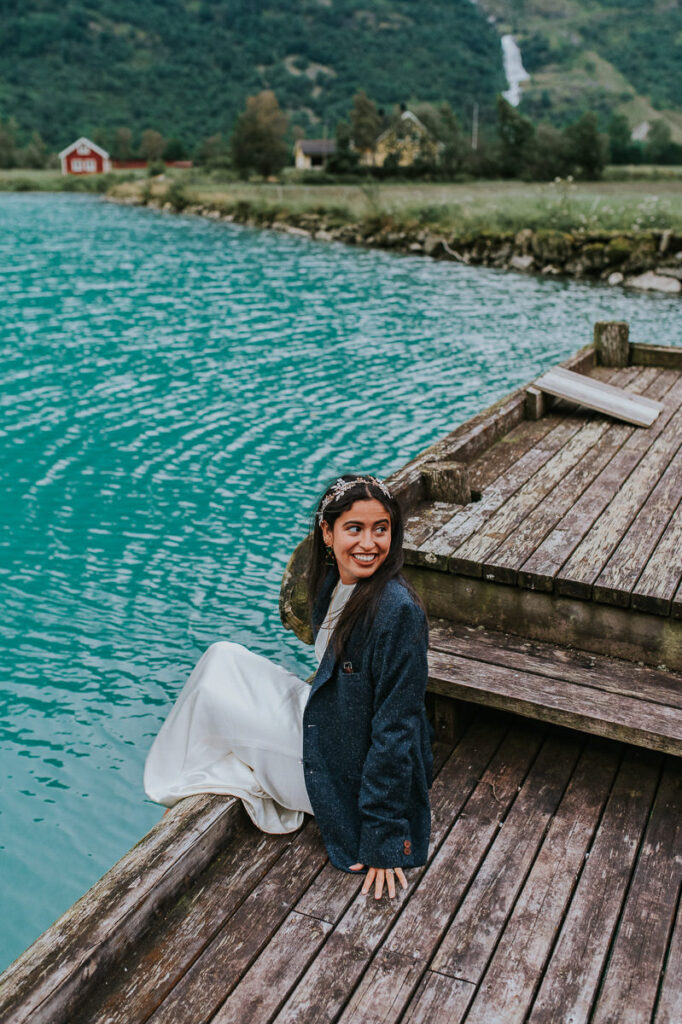 Beautiful bride sitting on a pier touching the water of the lake with her feet. She wears her husband's suit jacket and smiles