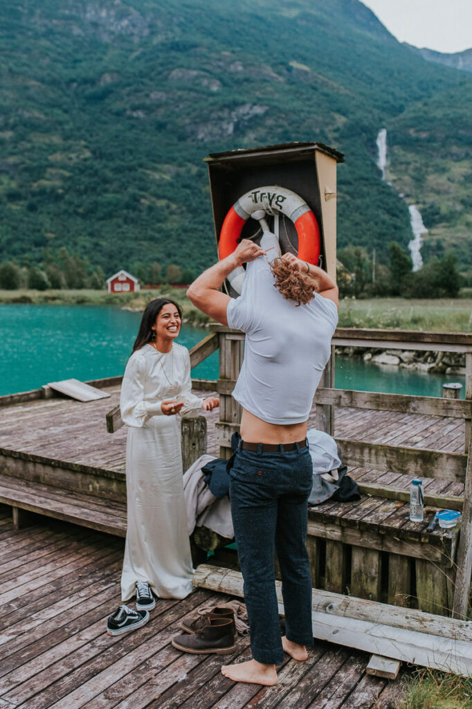 Groom is taking off his t-shirt while the bride giggles and is very excited. The groom is ready to jump in a beautiful glacier lake in Western Norway