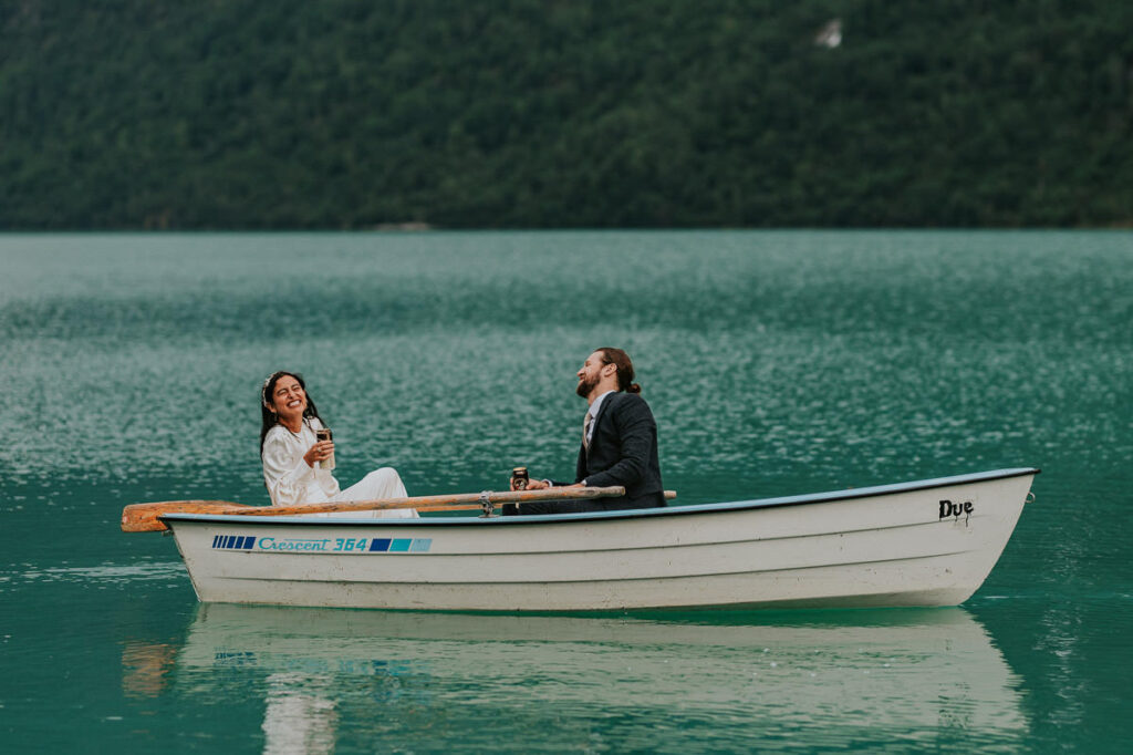 Birdal couple in a rowing boat drinks beers on a beautiful blue glacier lake Oldevatnet in Western Norway on the day of their intimate adventure wedding in Stryn