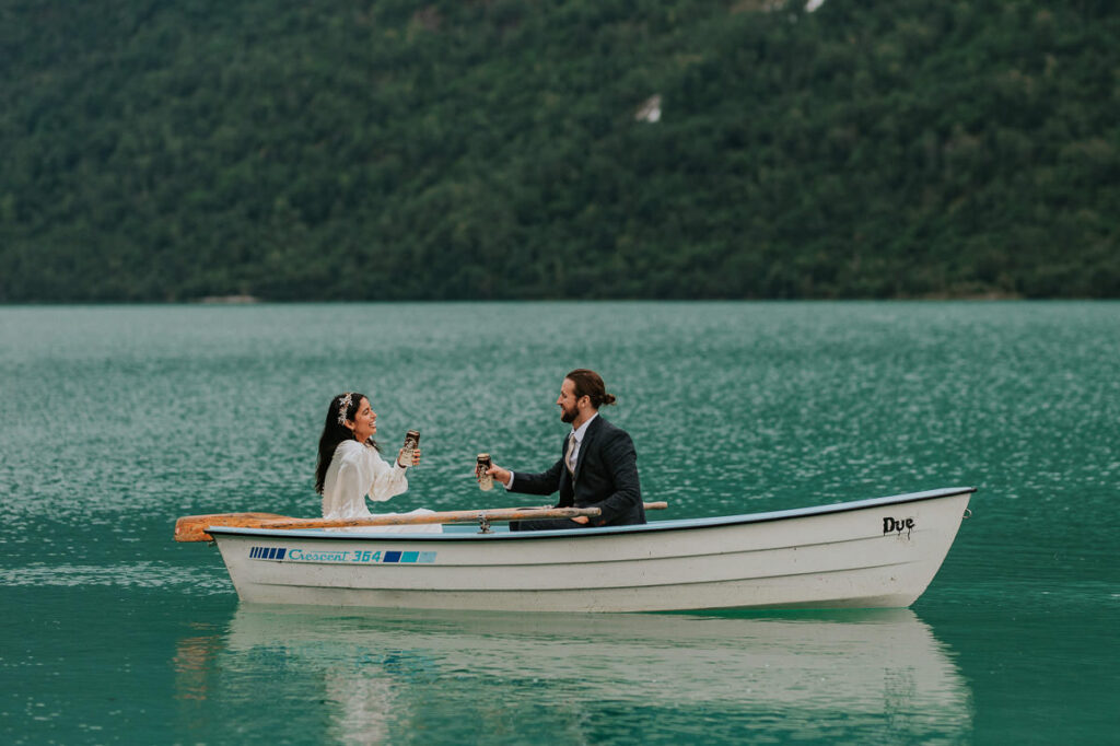 Birdal couple in a rowing boat drinks beers on a beautiful blue glacier lake Oldevatnet in Western Norway on the day of their intimate wedding 