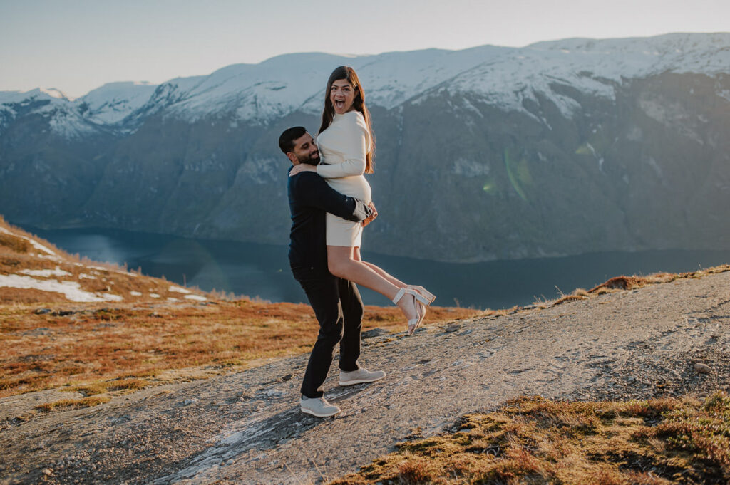 Happy couple on a mountaintop with snowy caped mountains in the background and a fjord view. The guy lifted his girlfriend and both look happy 