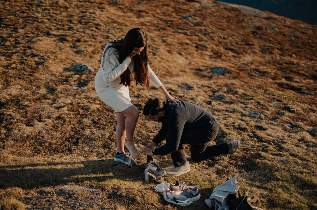 A guy is helping his girlfriend to change her shoes from high heels to sneakers for their mountain engagement photo session