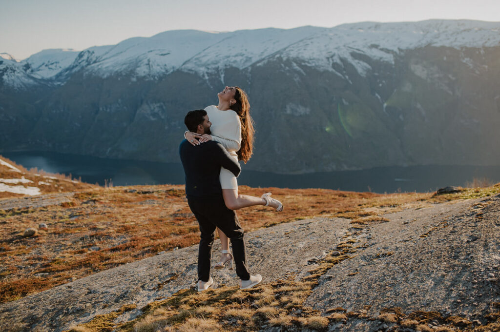 Happy couple on a mountaintop with snowy caped mountains in the background and a fjord view. The guy lifted his girlfriend and both look happy right after the proposal in Aurland Norway