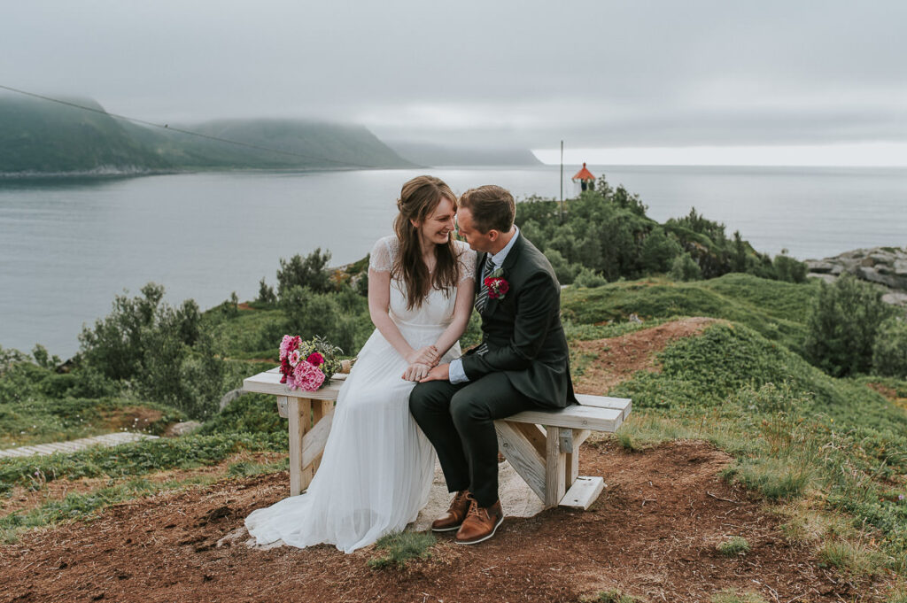 Bride and groom sitting on a wooden bench on Senja island with a lighthouse and foggy mountains in the background 