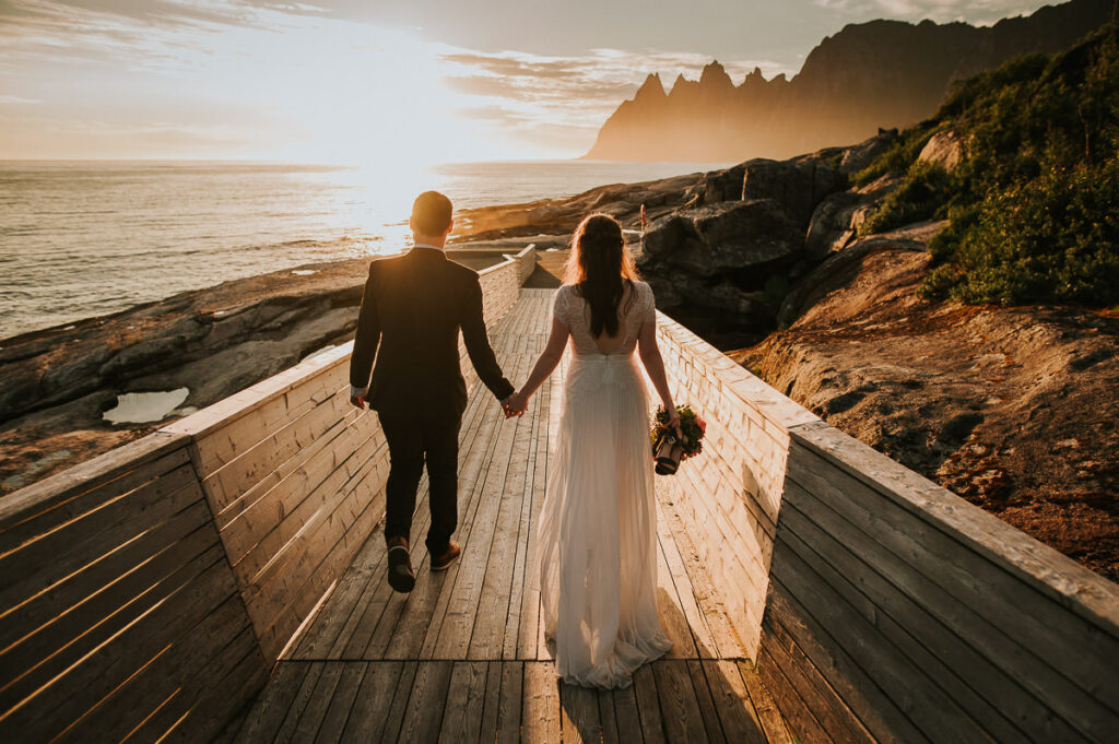 Bride and groom holding hands while walking towards spectacular mountains of Senja in the gorgeous midnight sun on their elopement day